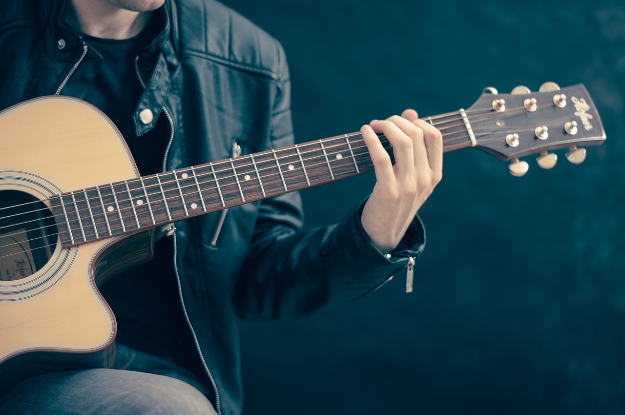 John Doe with a guitar sitting next to a brick wall with graffiti writing out democracy is a lie